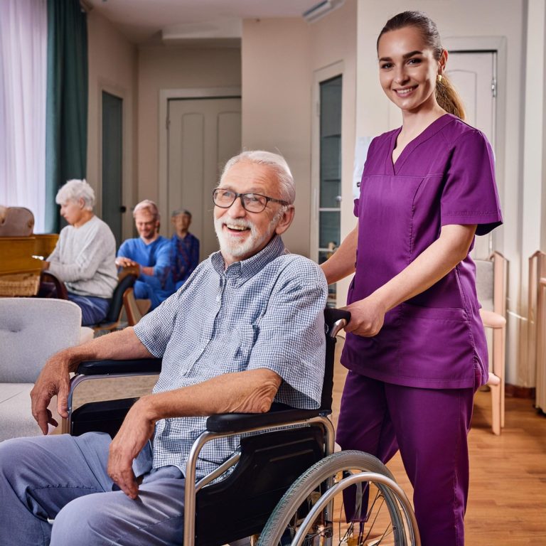 Lächelnder älterer Mann im Rollstuhl, unterstützt von einer Pflegerin von KRACH Arbeitnehmerüberlassung in lila Uniform.